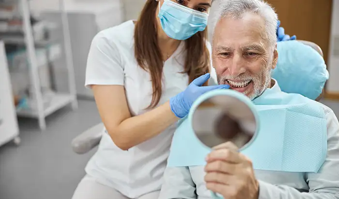 Older man sitting on dental chair and holding up a mirror to his new and restored smile with dental implants by Grins & Giggles Family Dentistry in Spokane Valley, WA